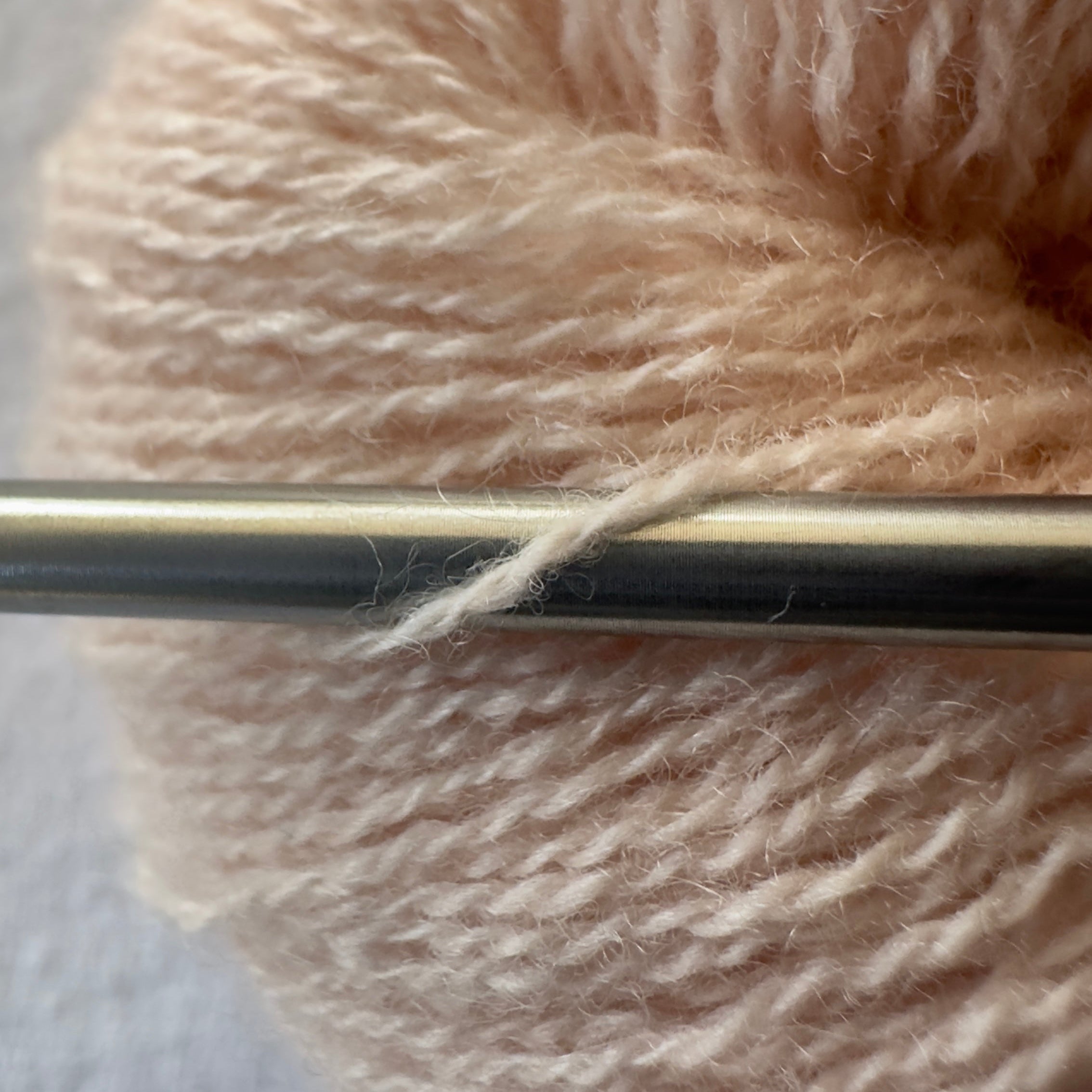 Close-up of Apricot Jam lace weight reclaimed cashmere yarn in a soft peach tone with delicate halo as it wraps around a sleek stainless steel knitting needle.
