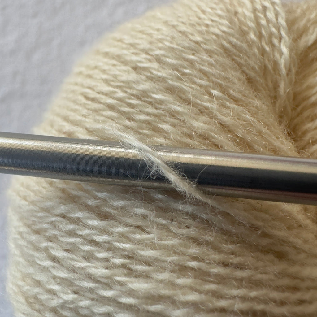 Close-up of lace weight reclaimed cashmere yarn in Raw Honey, a creamy yellow tone with soft halo as it wraps around a sleek stainless steel knitting needle.