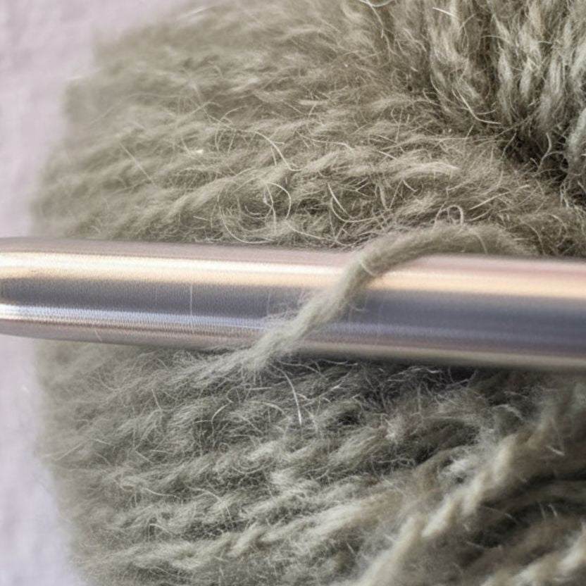 Close-up of reclaimed cashmere yarn strand wrapped around a smooth stainless steel knitting needle. The yarn is a soft, muted earthy green color with a luxurious halo of cashmere fibre against a textured white background.